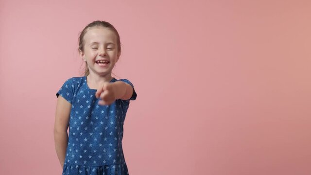 Beautiful cute girl points a finger and laughs. The concept of ridicule, ridicule. Blue dress with polka dots. The girl has long hair. Pink background.