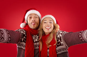 Silly young couple in Santa hats showing tongues while taking Christmas selfie on red background