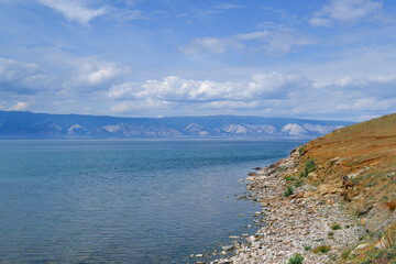 stone shore of lake baikal against the background of blue high mountains with clouds