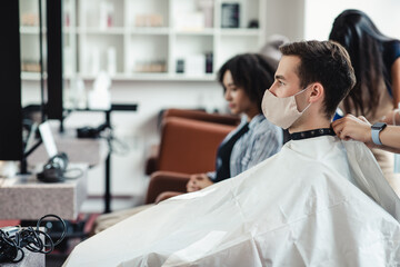 Young man in protective mask sitting in client chair at barber shop, waiting for haircut