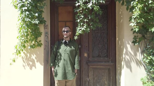 A man in sunglasses and a backpack behind his back stands in the doorway against the background of a wooden door. Vegetation on the building