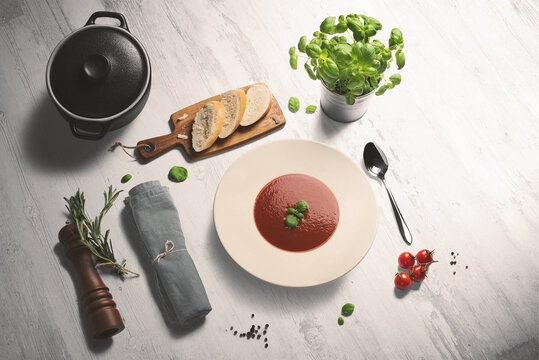 Bowl of fresh hot tomato soup on a whitewashed wooden counter. Fresh sliced bread, basil, cherry tomatoes and pepper kernels surround the bowl, accompanied by the black pot, napkin and pepper grinder.