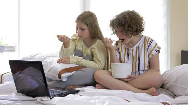 Two girlfriends of teenage age watching dramatic movie at home, sitting on the bed and eating ice cream. They are both moved and cry.