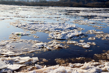 Ice drift on a river with blue high water and big water, white snow broken ice full of hummocks in it and sun reflection in sunny spring day.