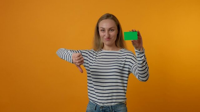 A young woman holds a blank card in her hand, looks at the camera and shows a finger down gesture. The concept of bad shopping, substandard product. Yellow background. 4K video.