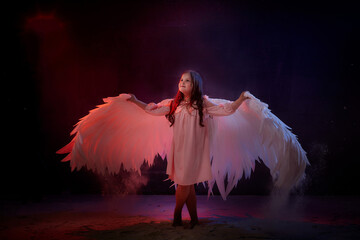 Small young model girl with white wings posing and dancing in dark black studo during photoshoot...