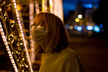 Obraz premium Portrait of a young woman in a yellow sweater with a coronavirus mask, against the background of yellow city lights at night. Bokeh in the background. Woman with sanitizer gel. Coronavirus concept.