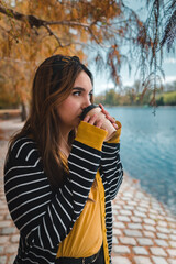 A young caucasian woman drinking coffee at autumnal park while looking at lake. Vertical