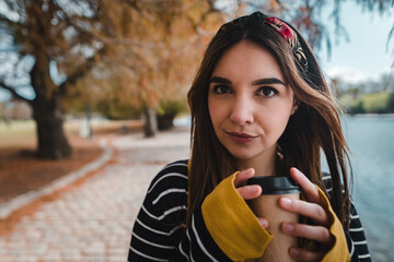 A pretty caucasian woman holding a cup of coffee and looking at camera at autumnal park