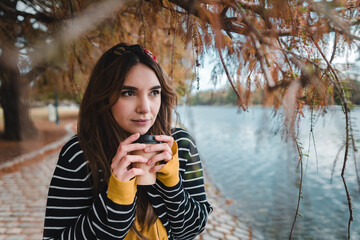 A pretty caucasian woman holding a coffee while standing below autumnal tree beside lake