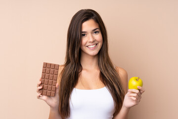 Teenager girl over isolated background taking a chocolate tablet in one hand and an apple in the...