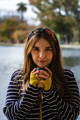 A pretty caucasian woman holding a cup of coffee while standing against a lake at park