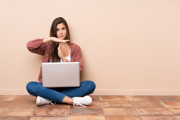 Teenager student girl sitting on the floor with a laptop making time out gesture