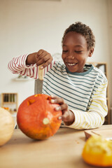 Vertical medium portrait of joyful young boy carving pumpkin with kitchen knife while preparing for...