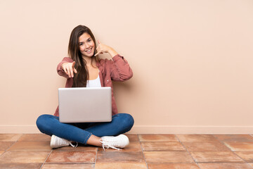 Teenager student girl sitting on the floor with a laptop making phone gesture and pointing front