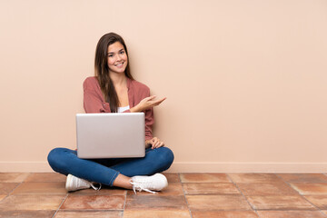 Teenager student girl sitting on the floor with a laptop presenting an idea while looking smiling towards