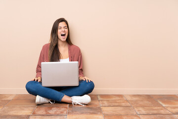 Teenager student girl sitting on the floor with a laptop shouting to the front with mouth wide open