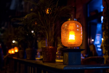 orange glass lantern decorated with cracks installed on the wooden railing of the terrace of a city restaurant with flowerpots, night scene illumination of a cafe, nobody.