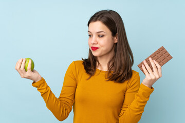 Teenager girl isolated on blue background having doubts while taking a chocolate tablet in one hand...