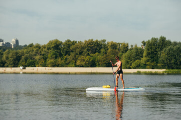 Athletic young man stands on a sup board on the river and paddles on a background of beach and...