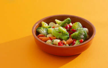 Frozen vegetables on the ceramic baking dish: paprika, carrot, broccoli, corn, tomato and green pea on the orange background. Healthy organic food.