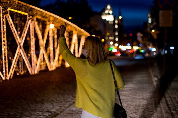 A young woman in a mask is catching a taxi. Woman in the city at night. Coronavirus concept. Woman wearing medical mask trying to catch a taxi in the city. Bokeh background. Health issues. Covid 19