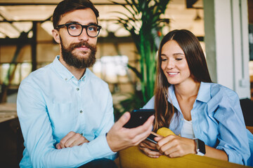 Portrait of Caucasian hipster guy in optical spectacles for eyes protection looking at camera during friendly meeting with carefree female, youthful man and woman blogging via cellphone device