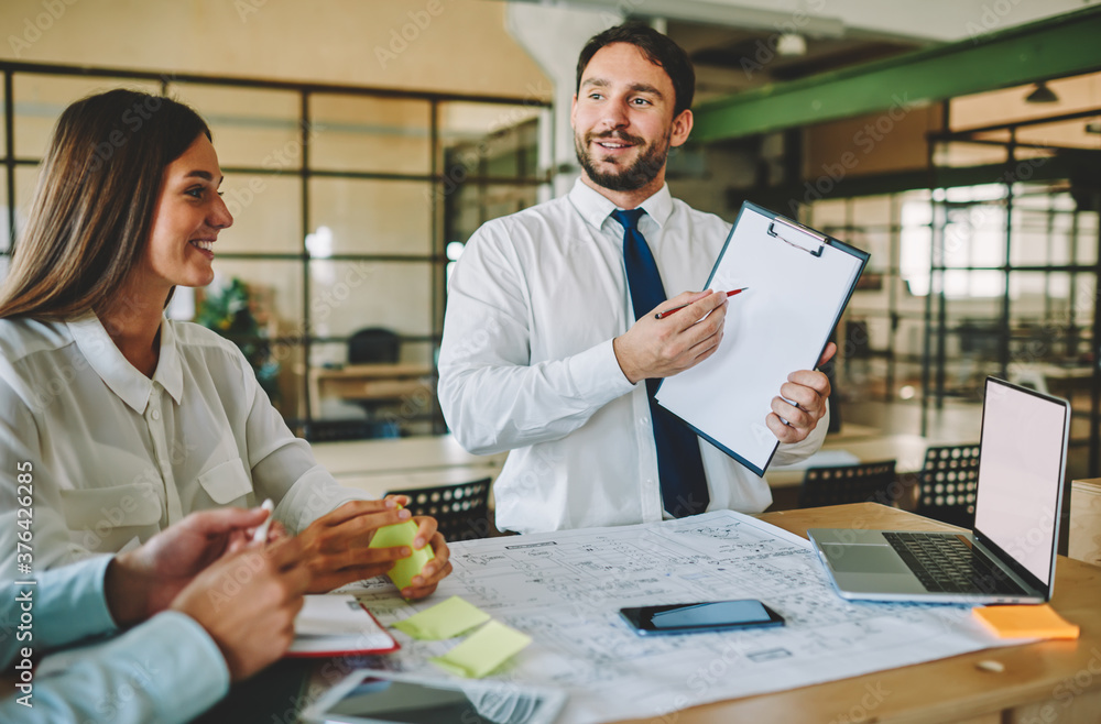 Poster smiling businessman showing paper folder to colleagues cooperating at desktop with blueprint and moc