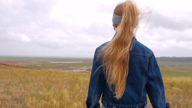 Adorable little girl in denim jacket back view on a hill with yellow grass field. Stylish child with long blonde hair on countryside cloudy sky landscape. Kid walking outdoor rural road trip faceless.
