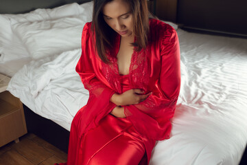 Irritable Bowel Syndrome or IBS. Asian woman in a silk nightgown and red robe suffering from period cramps while sitting on the bed in bedroom at night. Girls are not sleeping because of stomach pain.