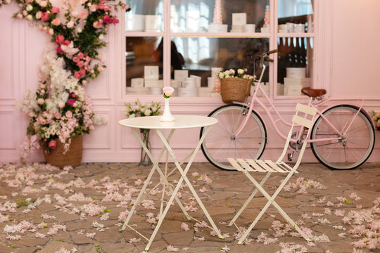 Empty cafe terrace with white table and chair. Pink exterior of the cafe restaurant. interior Street cafe. Cozy street with flowers and French-style cafe table. Decor facade of coffeehouse with bike. 