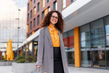 Frontal Pportrait of a smiling young woman with curly hair, wear in coat, at the modern building...
