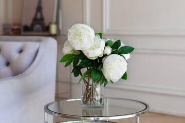 Vase with beautiful peony flowers on table near a grey sofa in living room. Cozy home decoration, fresh white peonies on coffee table in white room. Flowers in a vase at home. Scandinavia interior.	

