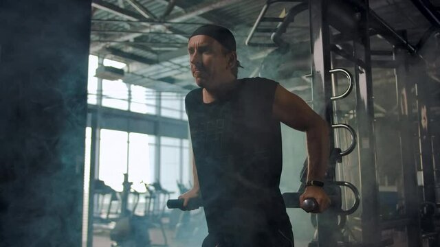 Mature man in the gym doing back push-ups on the simulator with parallel bars. Smoky gym in the background. Slow motion
