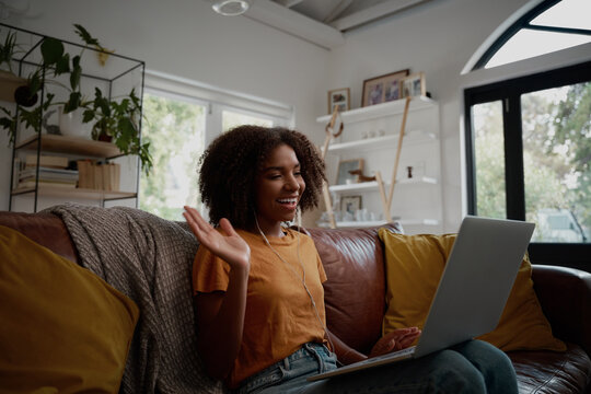 Smiling young african american woman relaxing on the couch waving hand while talking to friends on a video call at home - young blogger working from home