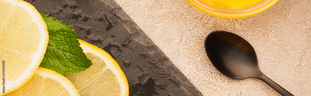 Poster top view of mint green leaves, lemon slices on black board near spoon on beige concrete surface, panoramic shot