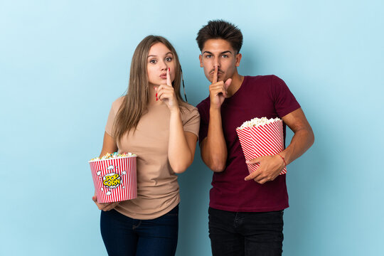 Couple eating popcorn while watching a movie isolated on purple background showing a sign of silence gesture putting finger in mouth