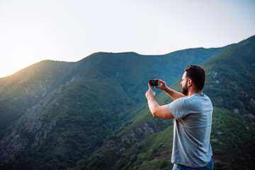 Young man taking a photo with his mobile phone in the mountainous landscape at sunset