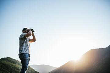 young man looking through binoculars on a mountain at sunset. Travel concept