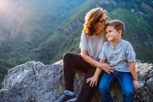 Mother kissing her six-year-old son sitting on the mountain at sunset