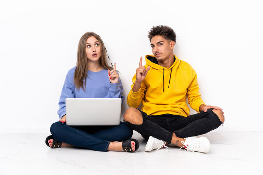Young couple with a laptop sitting on the floor isolated on white background thinking an idea pointing the finger up