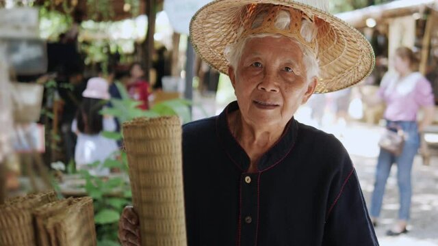 An elderly Asian female seller of a traditional Thai woven mat shop wearing local clothes , standing and wearing a traditional straw hat selling mats in front of the local store at the countryside.