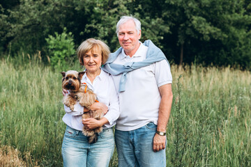 A mature couple is walking with a dog in a park. Elderly couple resting in nature with a dog. Close-up portrait of an elderly man and woman in white shirts and jeans. Stylish and modern grandparents.