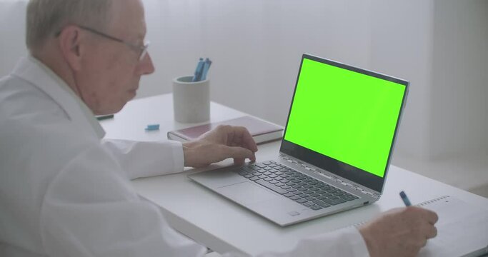 Elderly Male Doctor Is Reading Article In Internet, Looking At Chroma Key Display Of Laptop And Writing