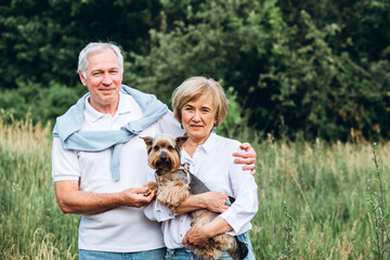 A mature couple is walking with a dog in a park. Elderly couple resting in nature with a dog. Close-up portrait of an elderly man and woman in white shirts and jeans. Stylish and modern grandparents.