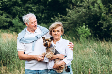 A mature couple is walking with a dog in a park. Elderly couple resting in nature with a dog. Close-up portrait of an elderly man and woman in white shirts and jeans. Stylish and modern grandparents.