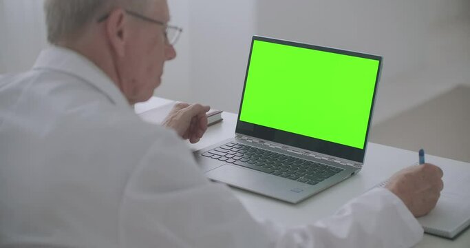 Laptop With Green Screen On Table Of Working Doctor, Man Is Looking At Display And Writing Notes, E-learning Concept