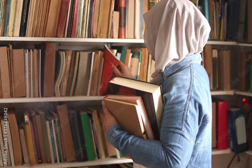 Wall mural Asian muslim woman wearing hijab picking book in bookshelf, education concept, reading learning studying in library