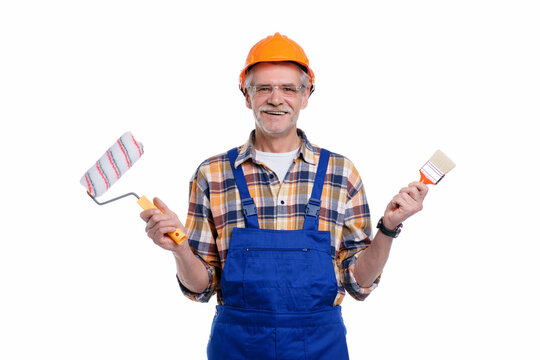 Photo of positive senior male builder holding paint roller and brush in hand, wearing special uniform, apron and protective helmet, isolated on white background