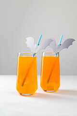 Halloween Mocktails. Two glasses of orange non-alcoholic cocktail for Halloween party on white table. Straws are decorated with cut paper bats. Selective focus, copy space, vertical orientation.